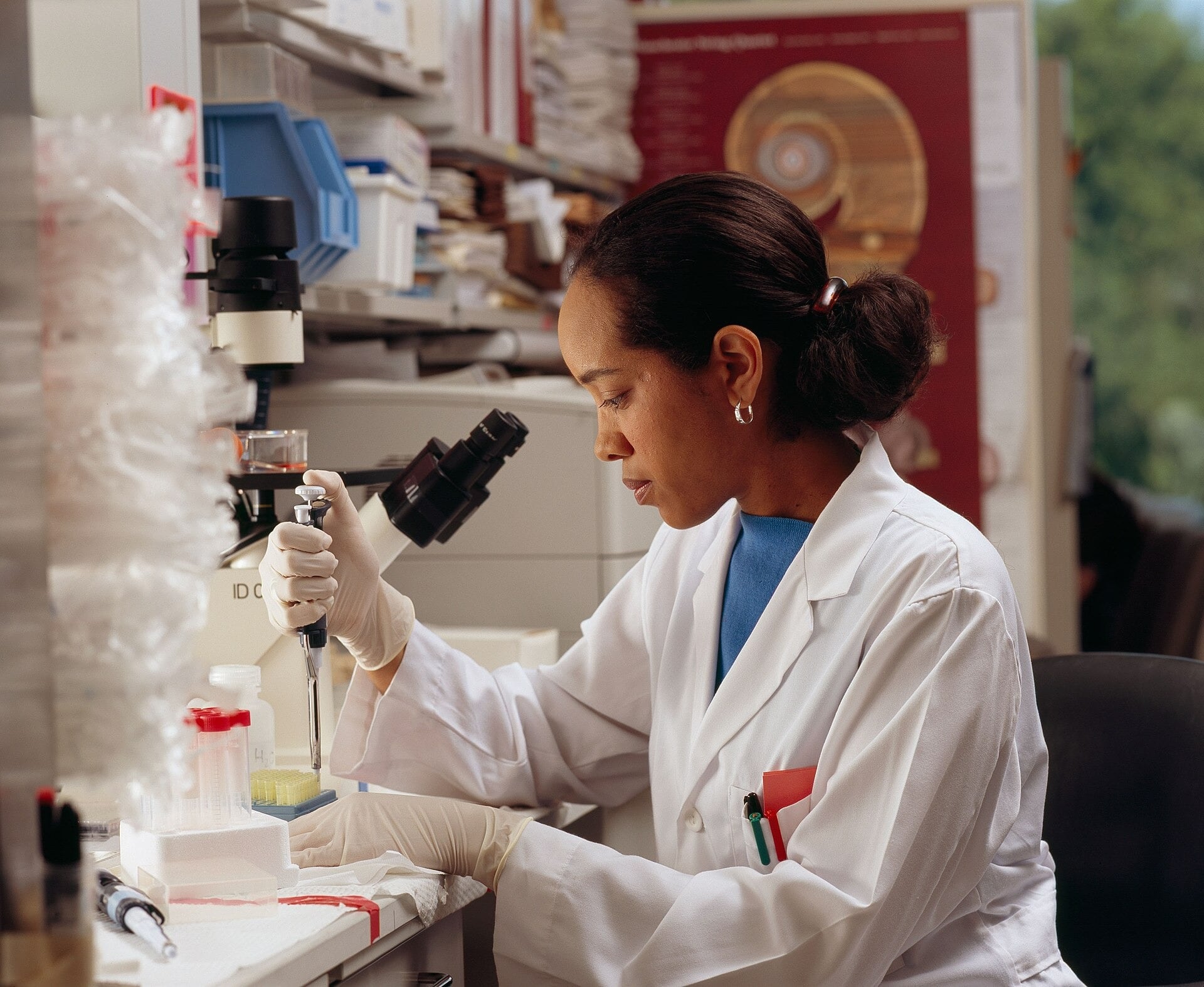 A researcher in a lab coat and gloves uses a pipette at a laboratory bench