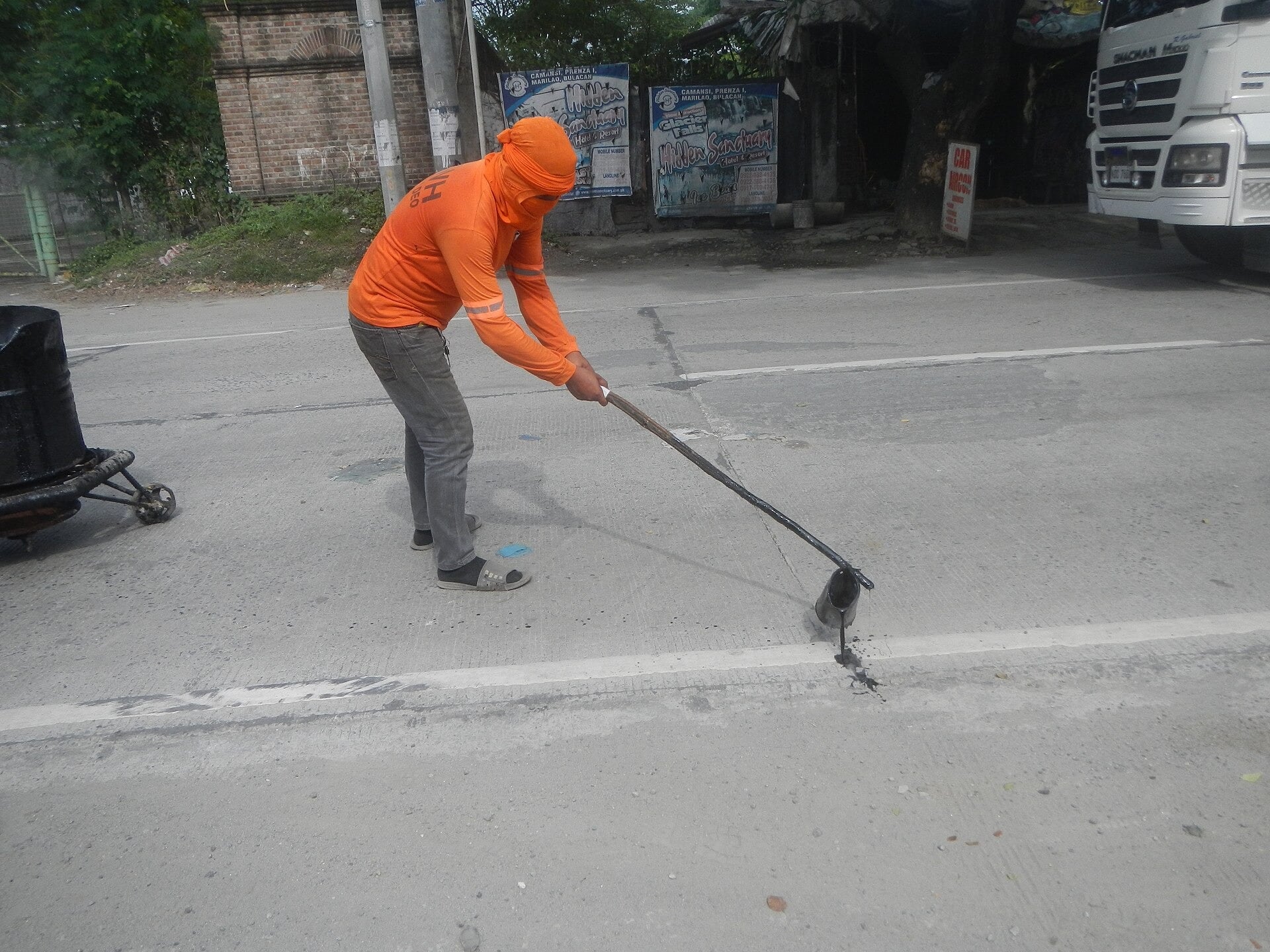 A road worker in an orange hooded shirt spreads hot asphalt along a road seam with a long-handled tool