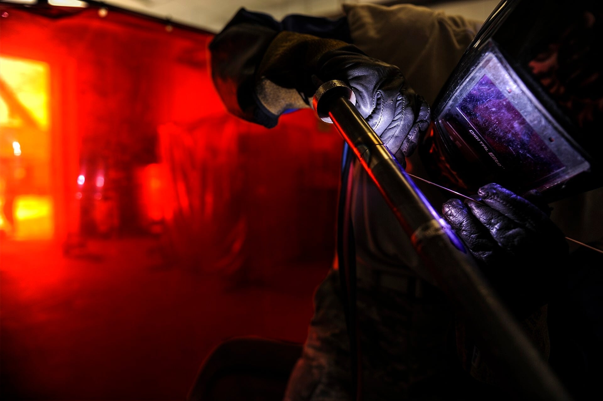 A welder wearing a protective helmet and gloves works on metal with a glowing tungsten arc