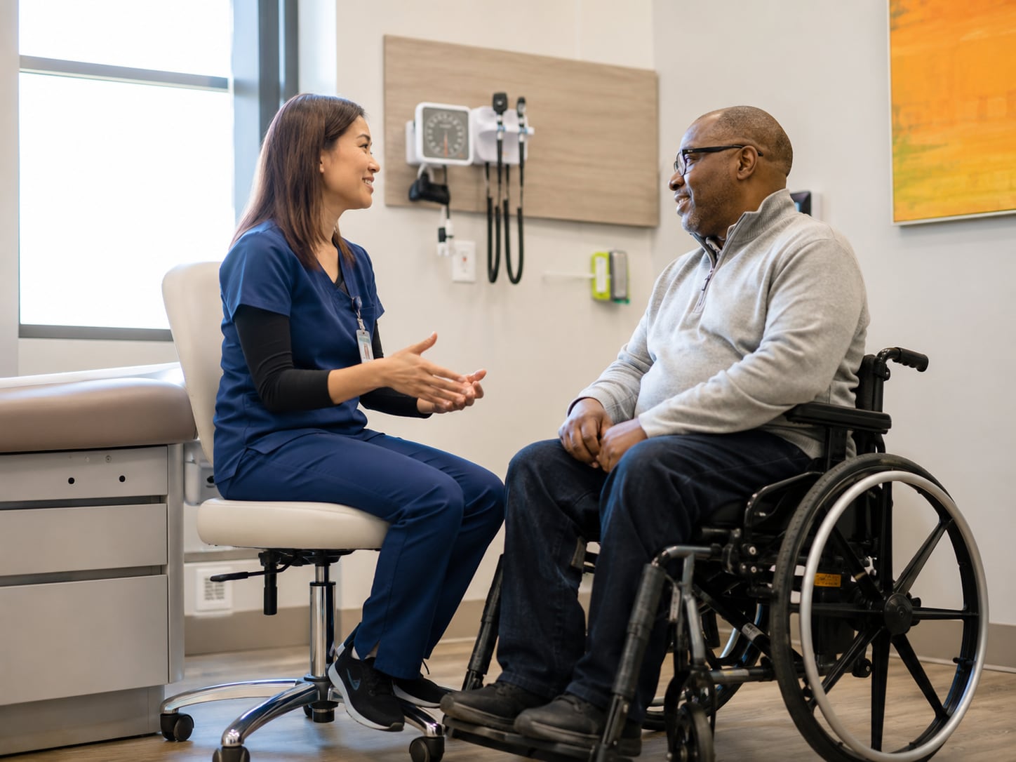 A female nurse talks with an older Black man seated in a wheelchair in an exam room