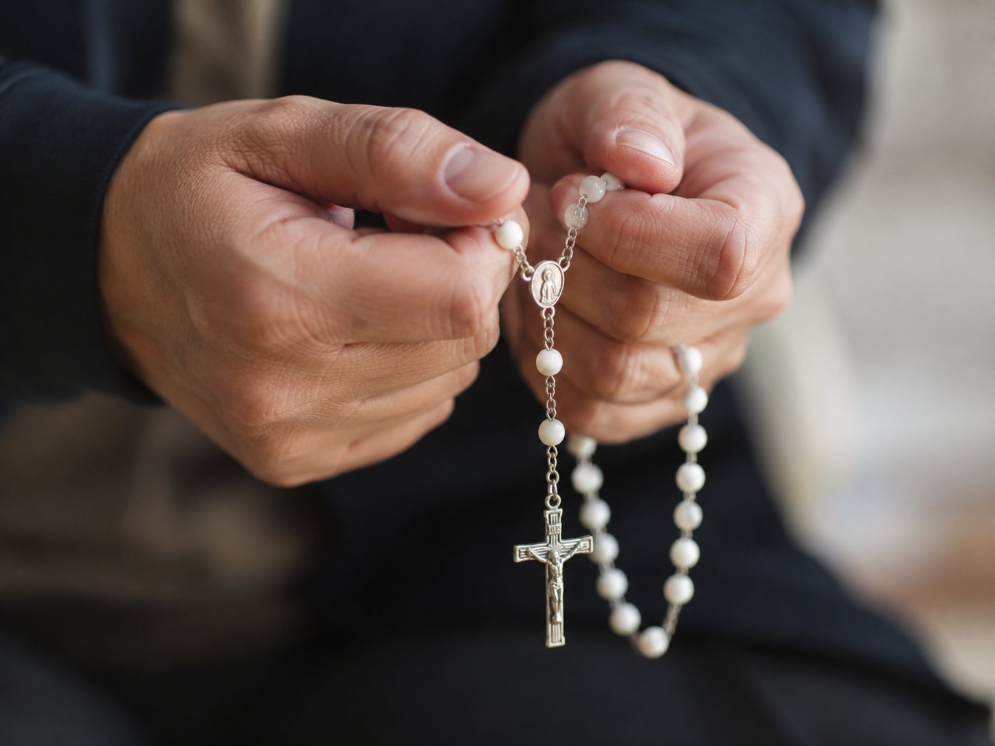 Two hands hold a pearl rosary with a silver crucifix against a dark sweater