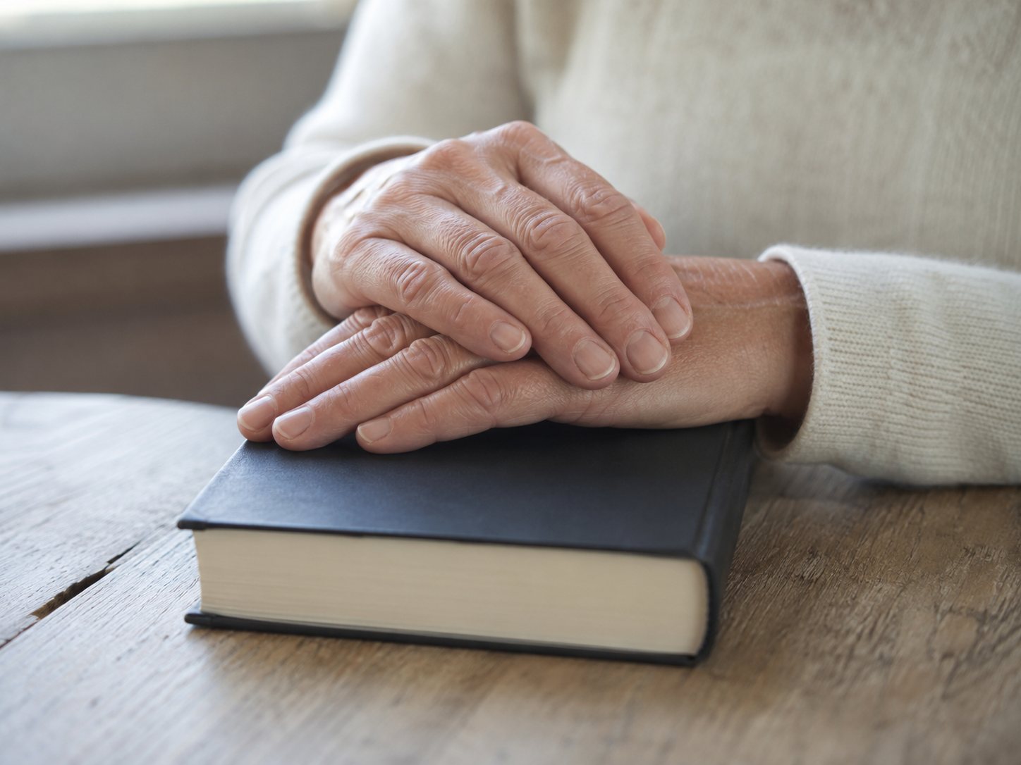 Two older hands rest one over the other on a closed hardback book on a wooden table