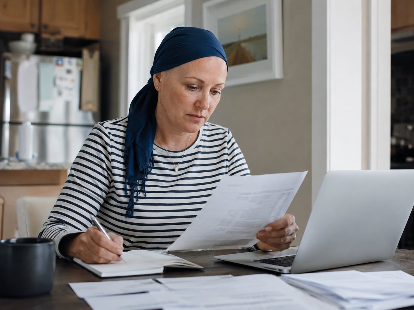 A woman in headscarf stands in a kitchen reading a letter or paper