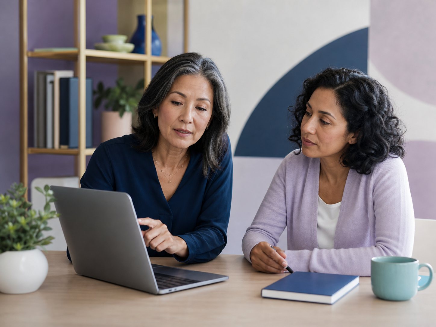Two women sit side by side reviewing a laptop and a notebook, comparing versions of a text.