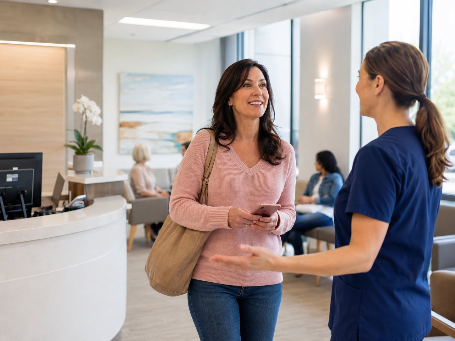 Woman with a shoulder bag smiles while talking with a staff member in scrubs in a hospital lobby.