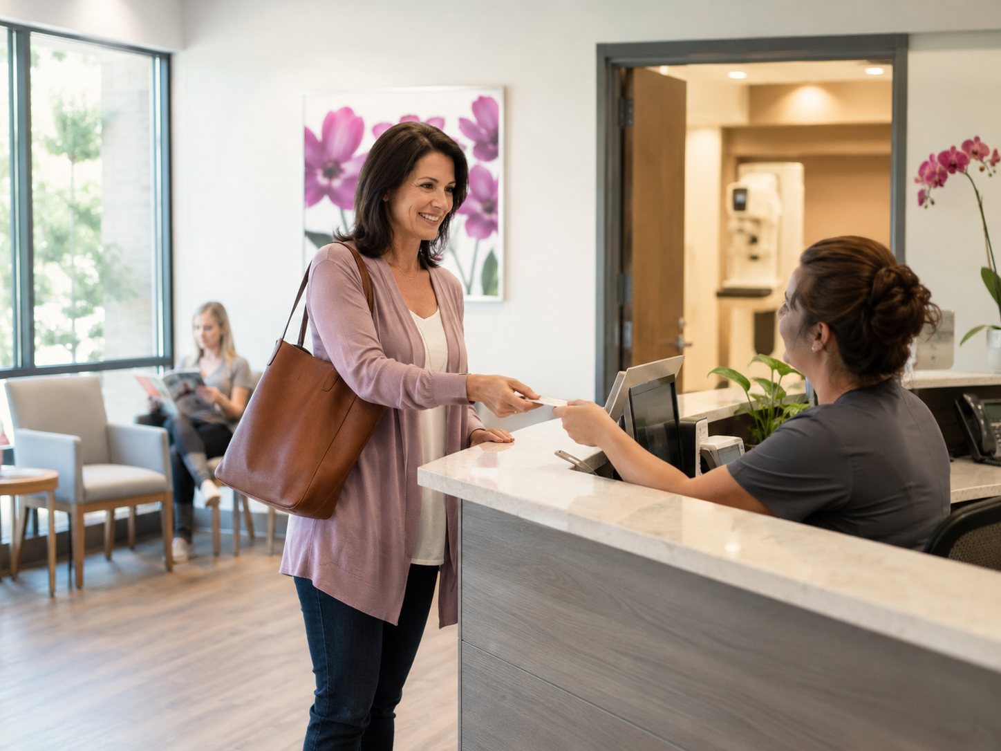 Woman with a shoulder bag hands a card to a receptionist in scrubs at a clinic front desk.