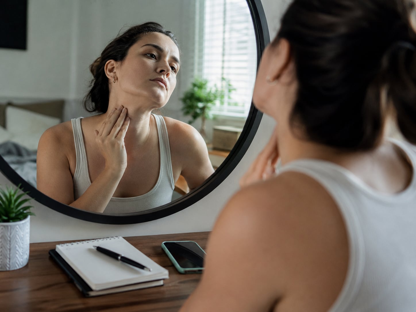 A woman examines her own neck in a bathroom mirror