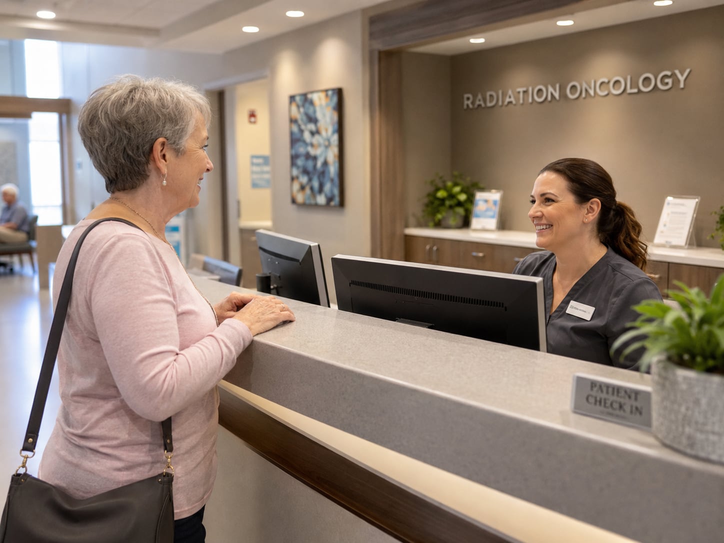 An older woman with a shoulder bag stands at a reception counter speaking with a seated staff member, beneath lettering reading “Radiation Oncology”.