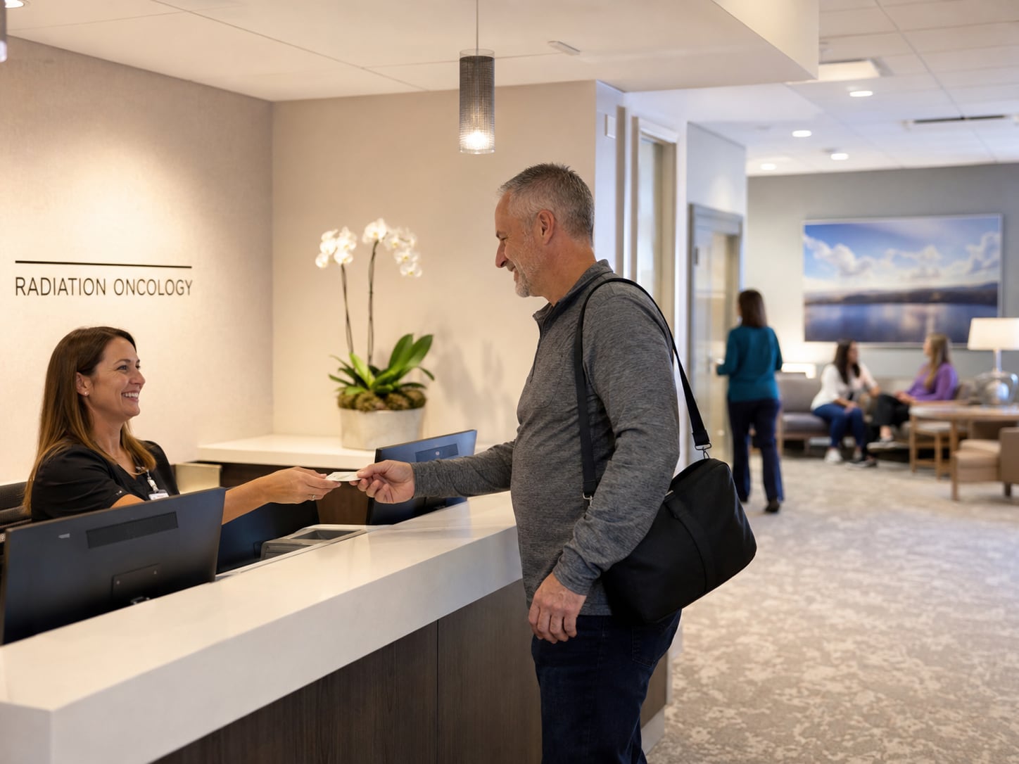 A man with a shoulder bag hands a card to a receptionist at a front desk beneath a wall sign reading “Radiation Oncology”.