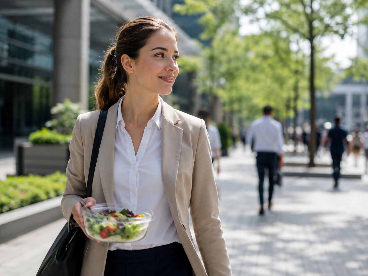 Woman in a blazer walks a tree-lined city sidewalk carrying a clear bowl of salad.