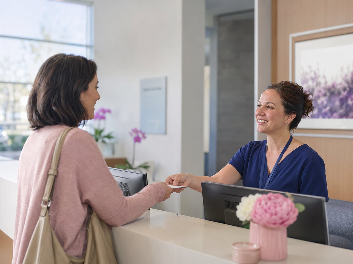 A woman greets a smiling clinician at a reception counter