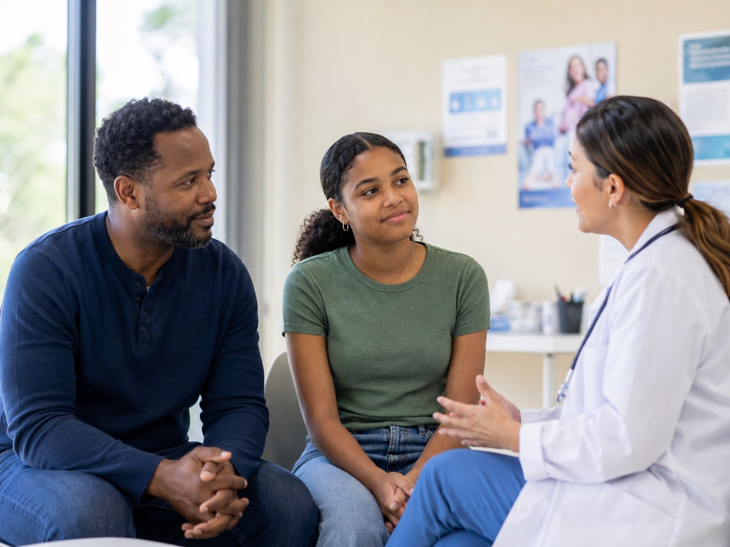 A man and teenage girl talk with a doctor holding a tablet in an exam room