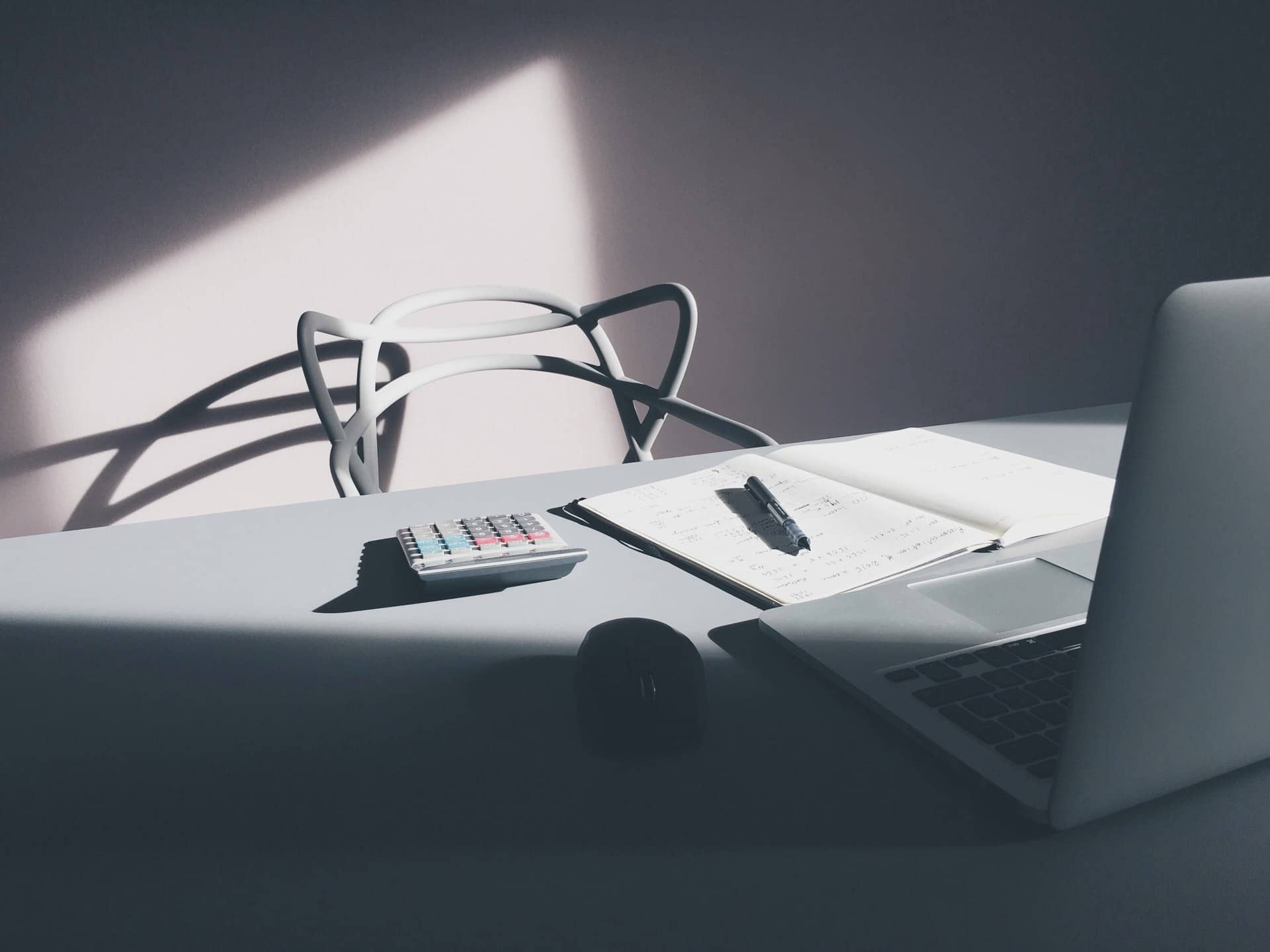 A desk with a calculator, an open notebook with a pen, and a laptop in side light.