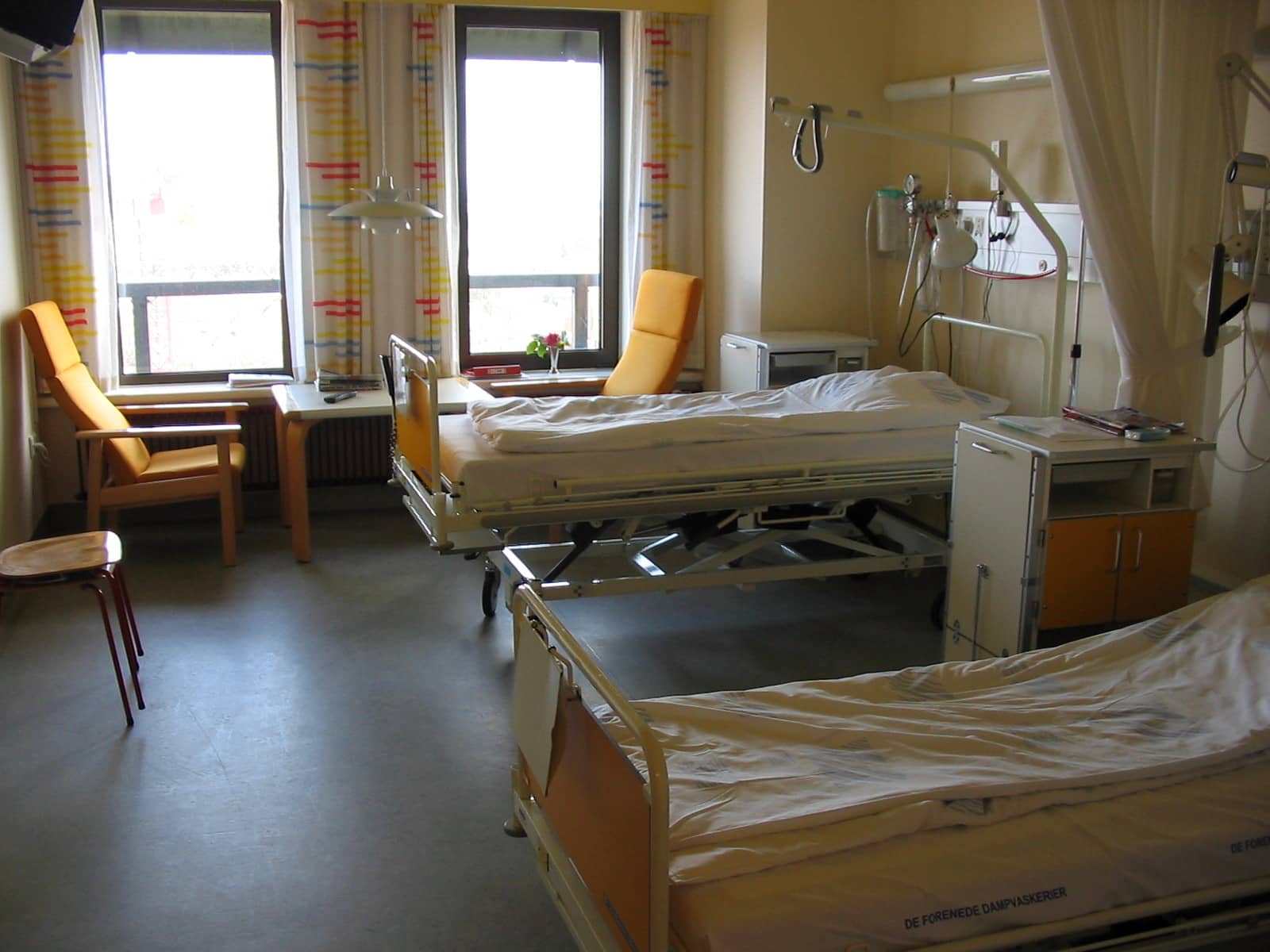A hospital room with two empty beds, two armchairs and large windows.