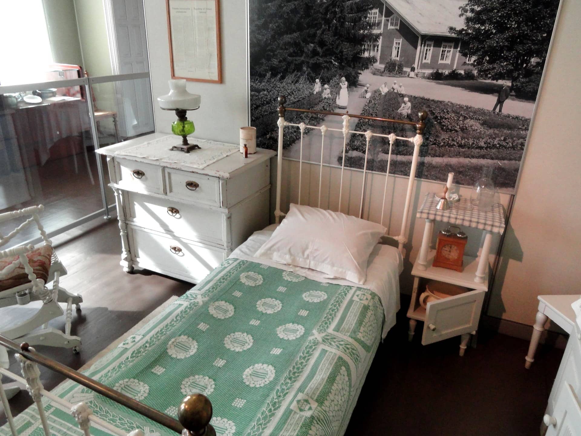 A museum display of an old patient room with an iron bed, green coverlet and oil lamp.