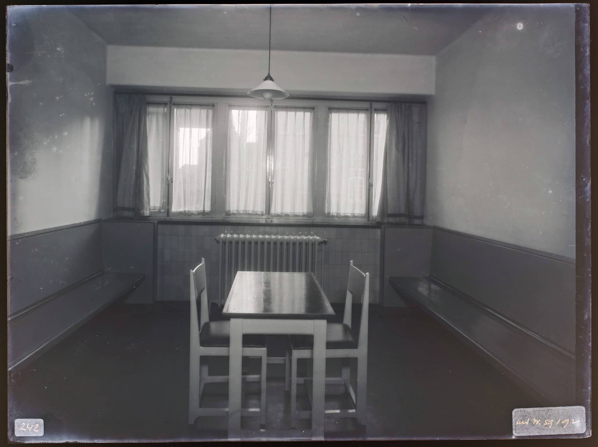 A black-and-white photo of an empty waiting room with a table, two chairs and wall benches.