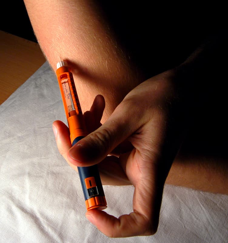 A hand holding an orange and blue injection pen against the skin of an upper arm.