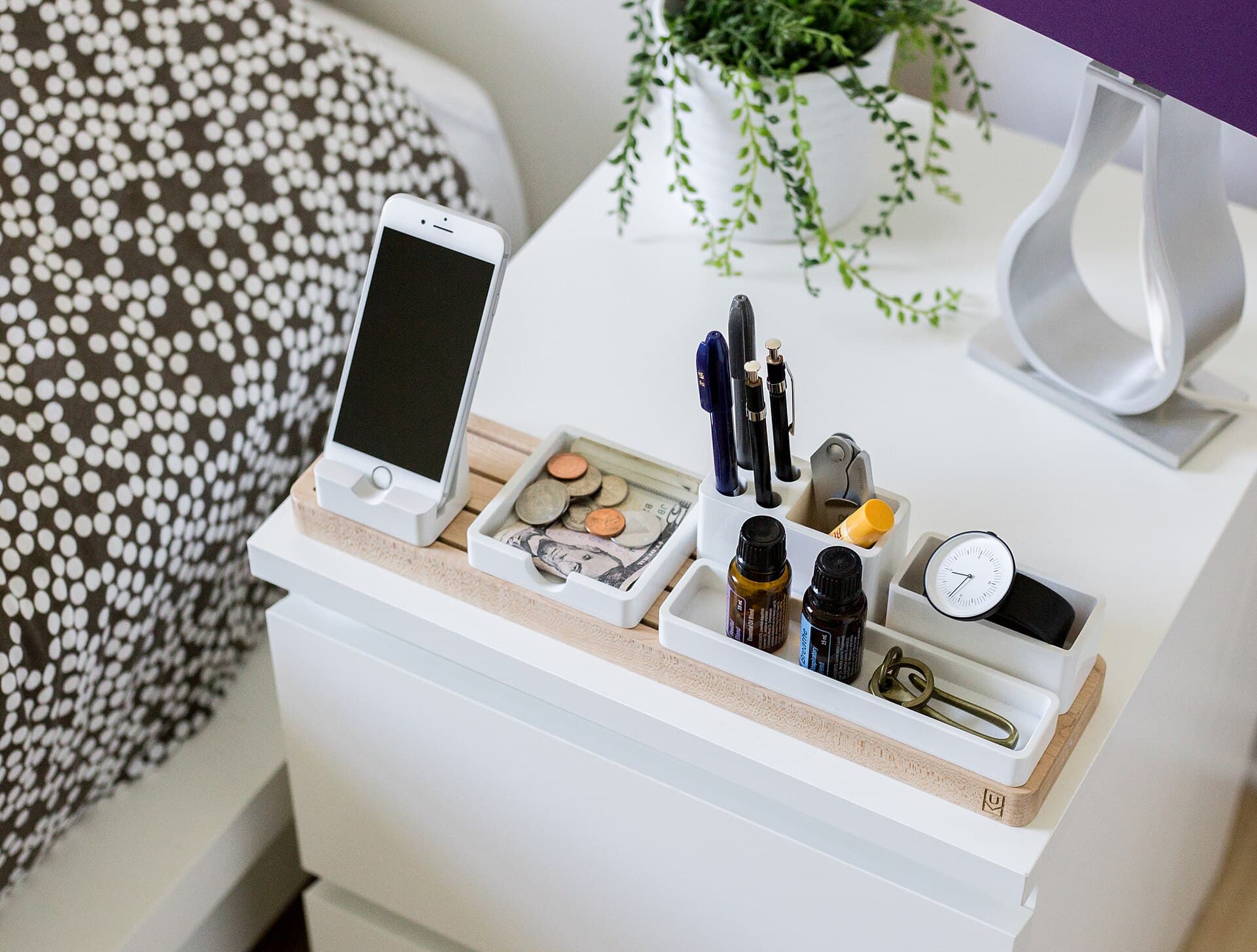 A smartphone standing in a dock on a white bedside table beside a patterned bed, with pens, coins, a watch and a plant.