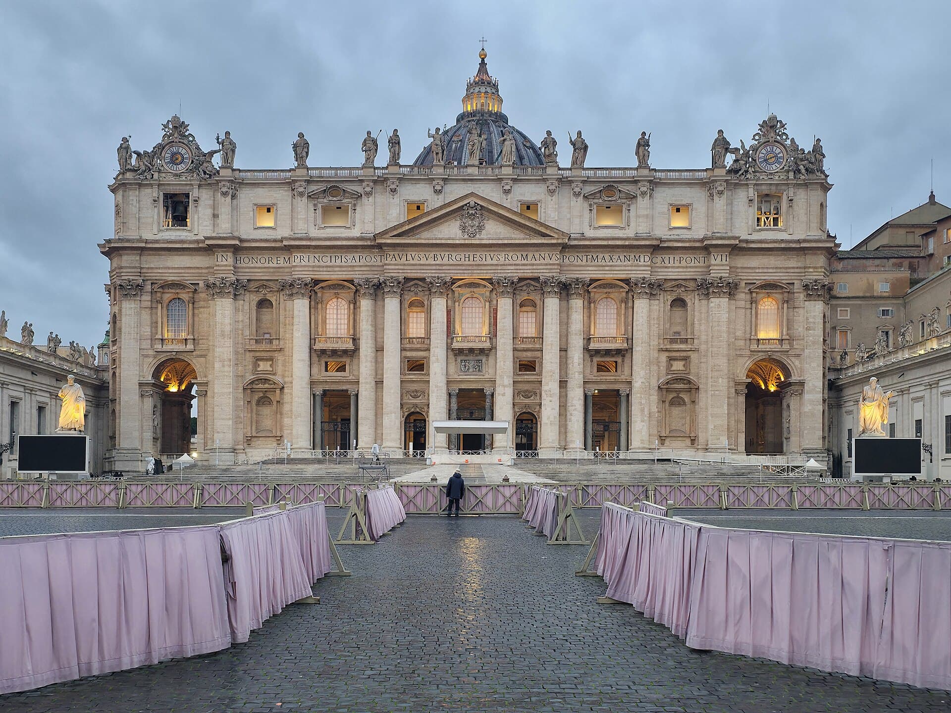 Front facade and dome of St. Peter's Basilica in Rome at dusk, with a cobbled approach and pink crowd barriers.