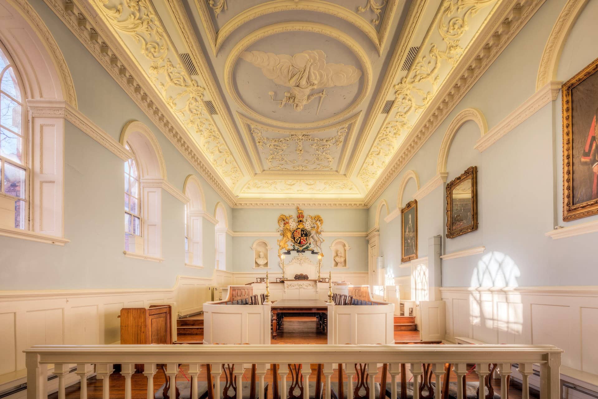 Empty historic courtroom with a decorated plaster ceiling, wooden benches and a royal coat of arms.
