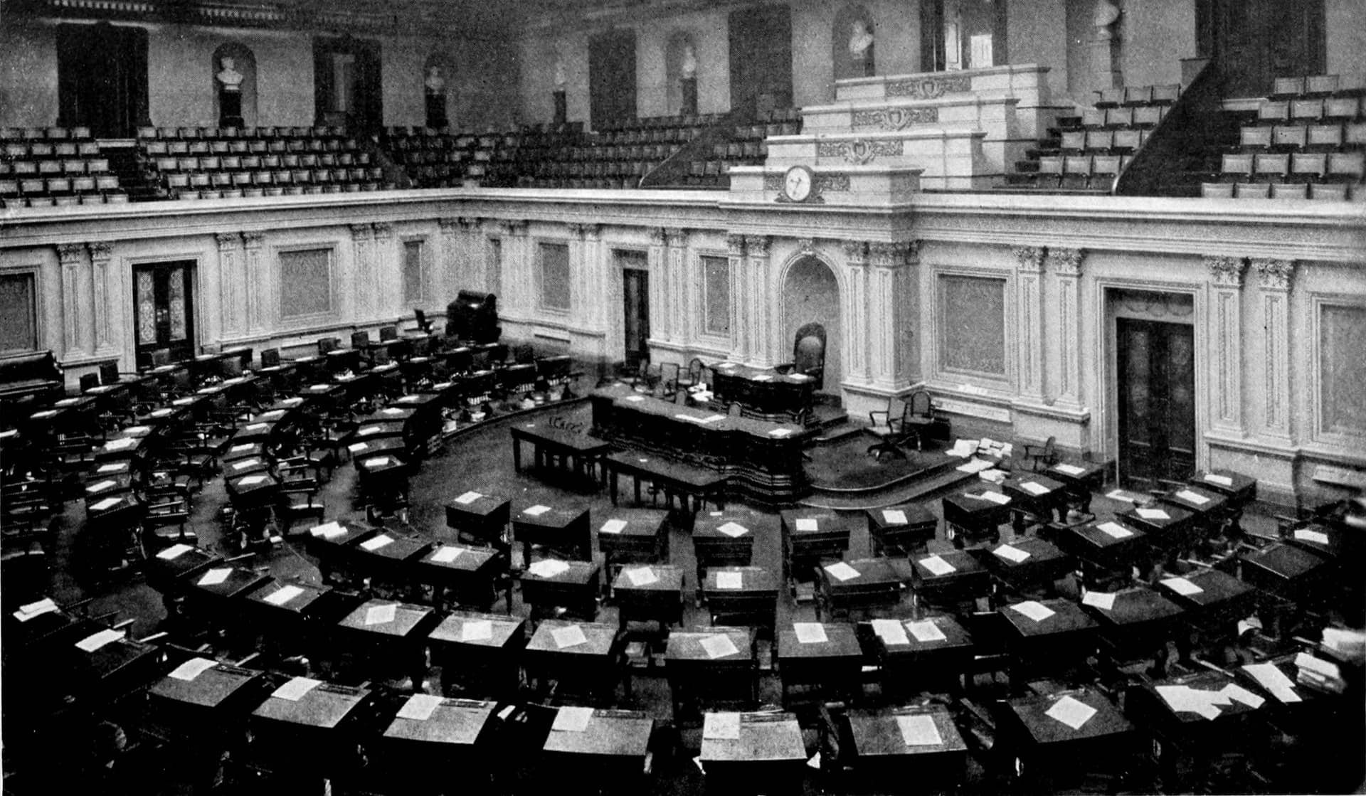Black-and-white photograph of an empty legislative chamber with rows of desks.