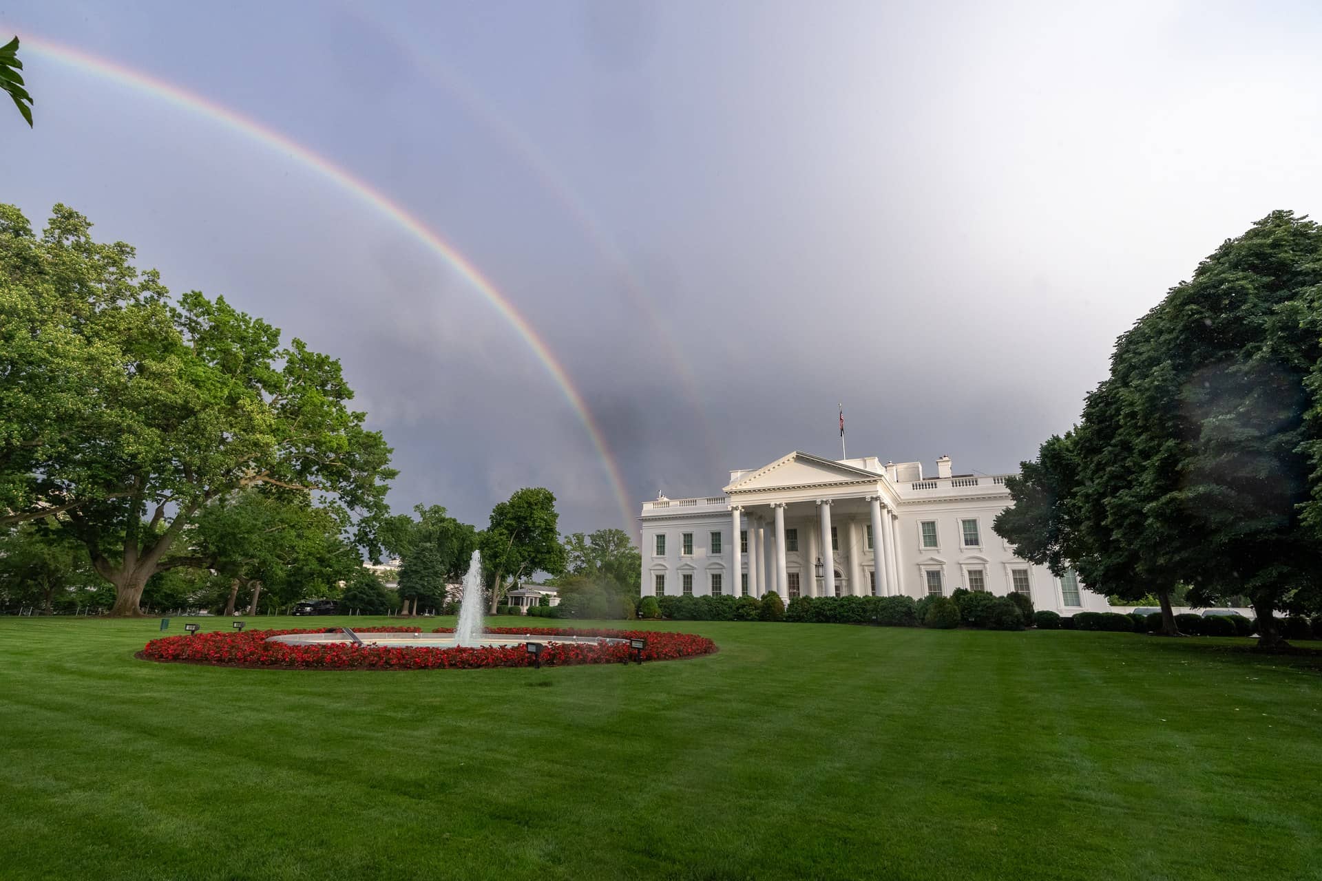 A large white columned building beyond a lawn and fountain, with a rainbow in a cloudy sky.