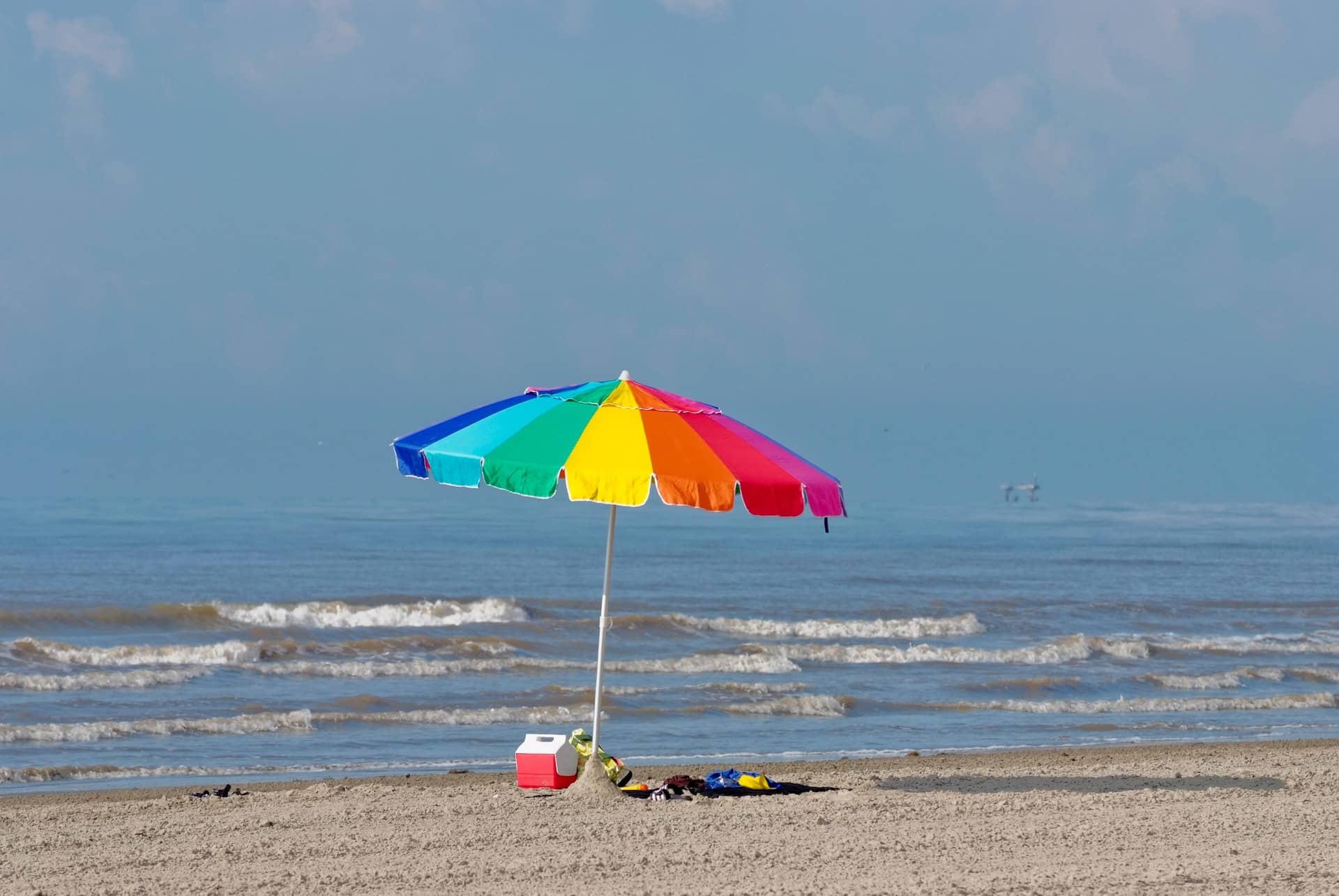 A rainbow-coloured beach umbrella standing on a sandy shore beside the sea.