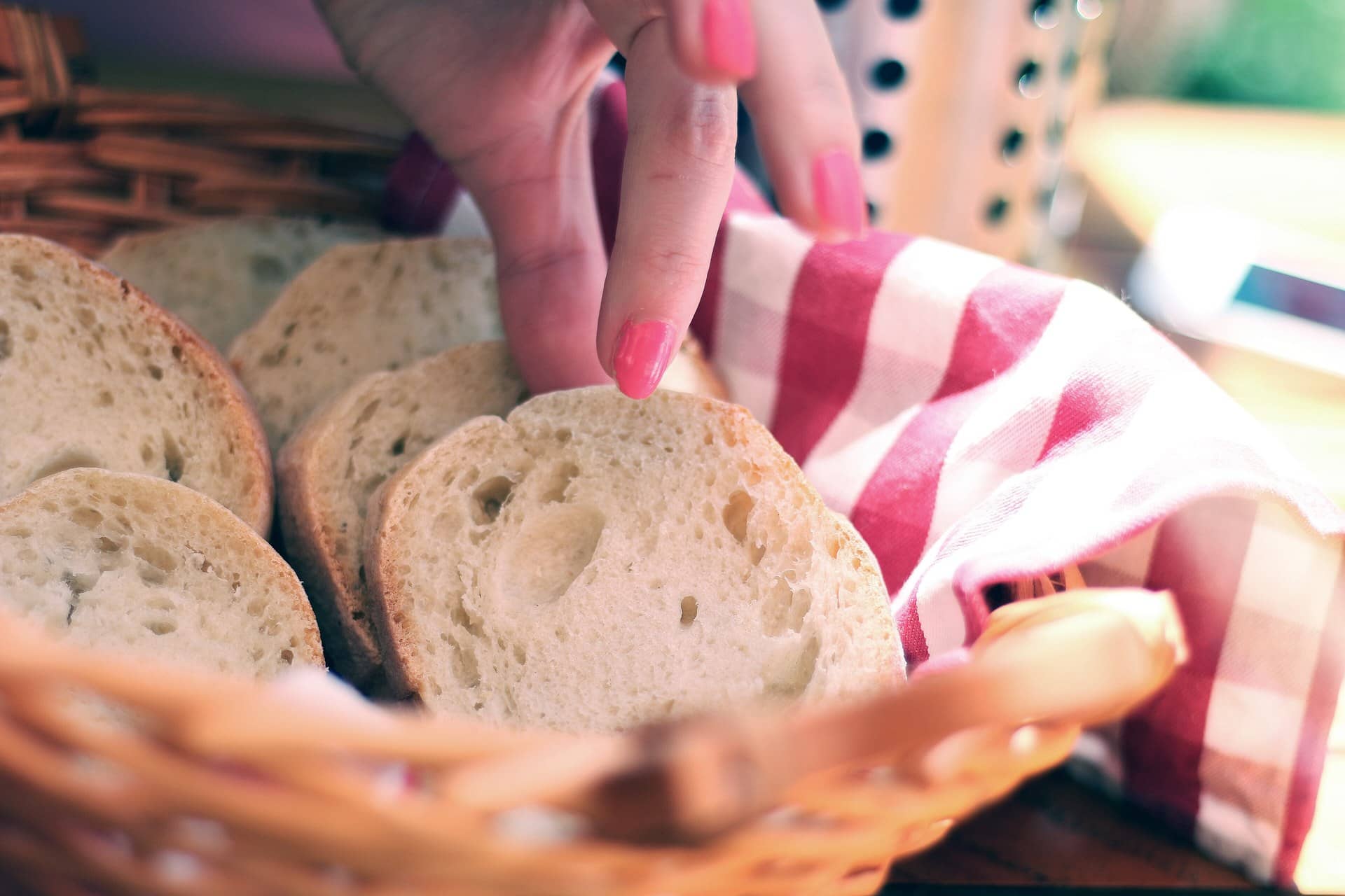 Slices of baguette in a wicker basket with a checked cloth and a hand reaching toward a slice.