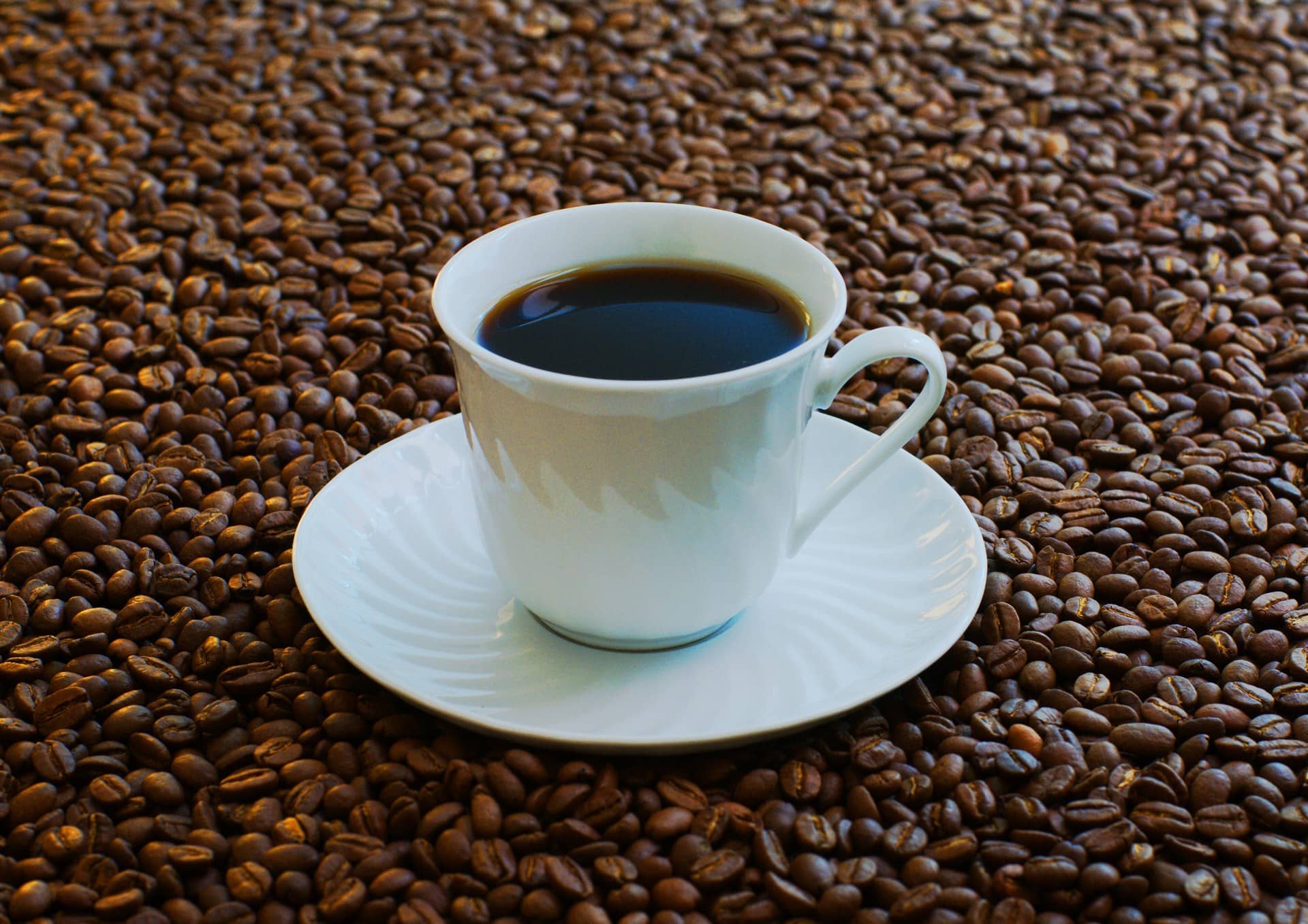 A white cup of black coffee on a saucer surrounded by roasted coffee beans.