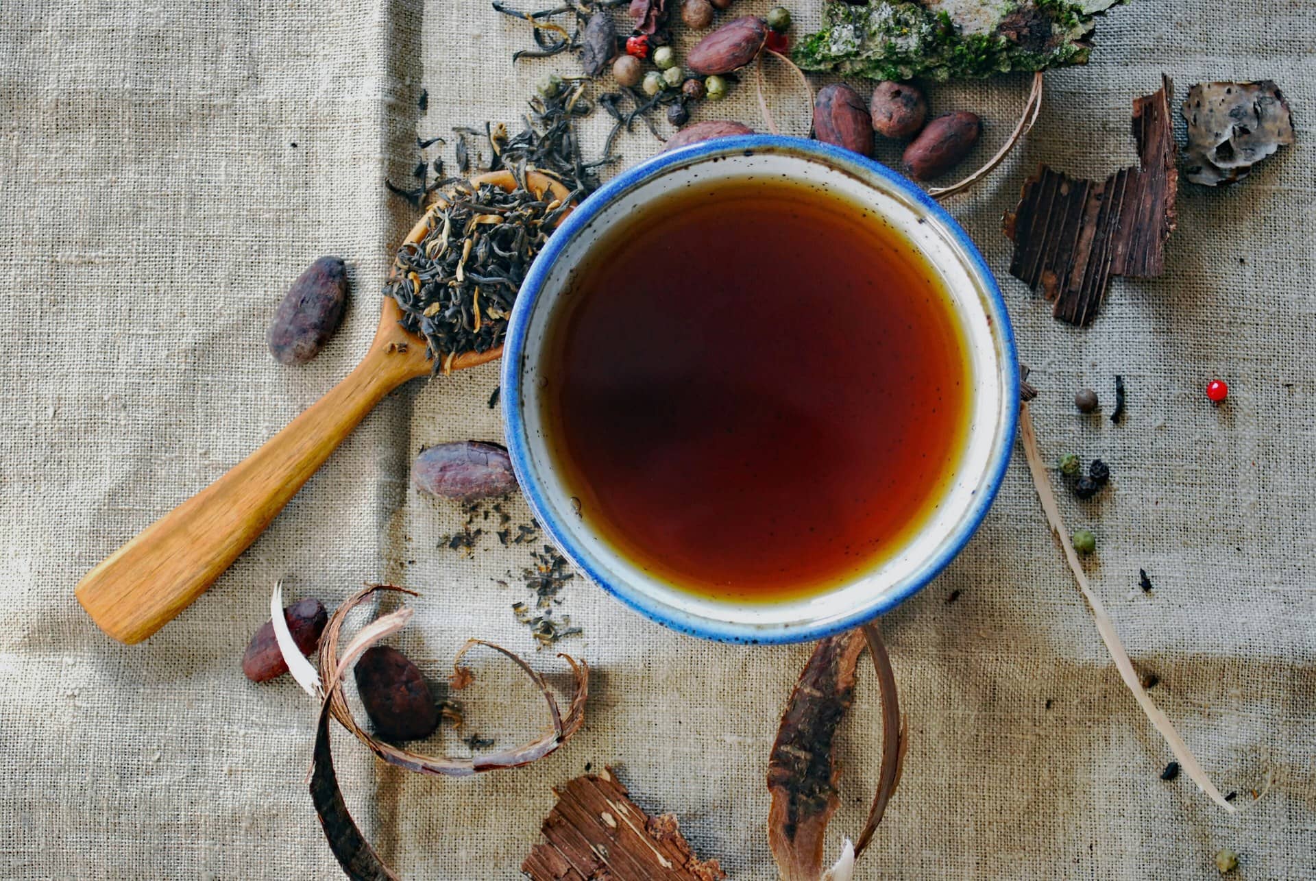 A bowl of dark tea with loose leaves, spices, bark and a wooden spoon on linen.