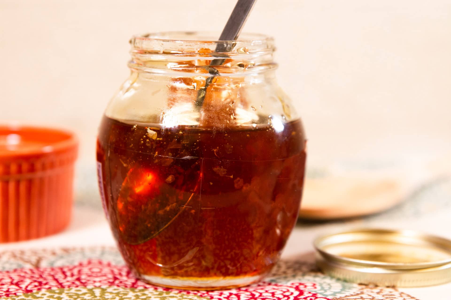 A glass jar of dark amber syrup, likely honey, with a spoon in it on a patterned cloth.
