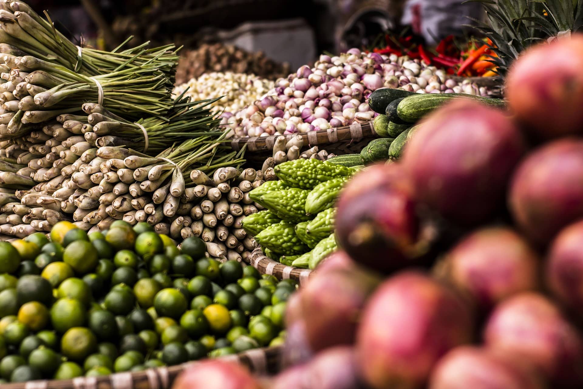 A market stall piled with lemongrass, limes, onions and other produce, with out-of-focus onions in front.