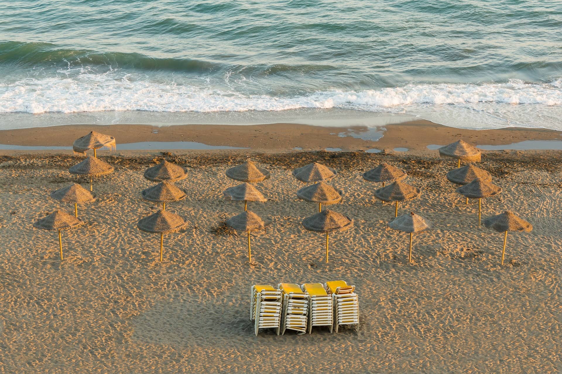 Rows of straw parasols on a sandy beach beside the sea, with stacked sun loungers.