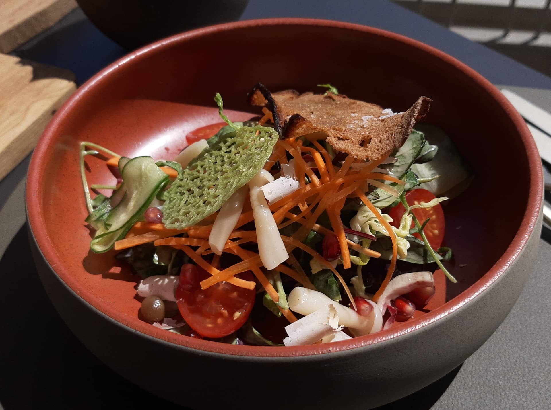 A bowl of salad with carrot, tomato, cucumber and a toasted bread crisp.