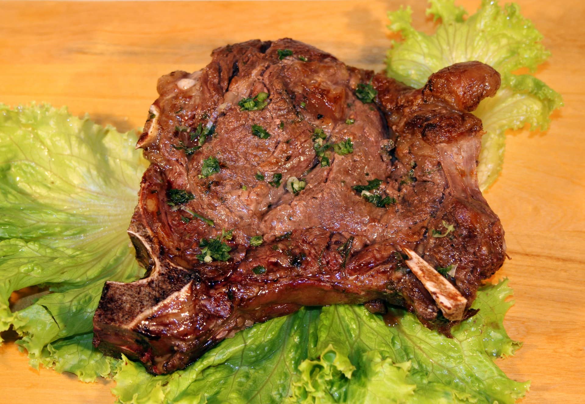 A cooked bone-in cut of meat with herbs on lettuce leaves on a wooden board.