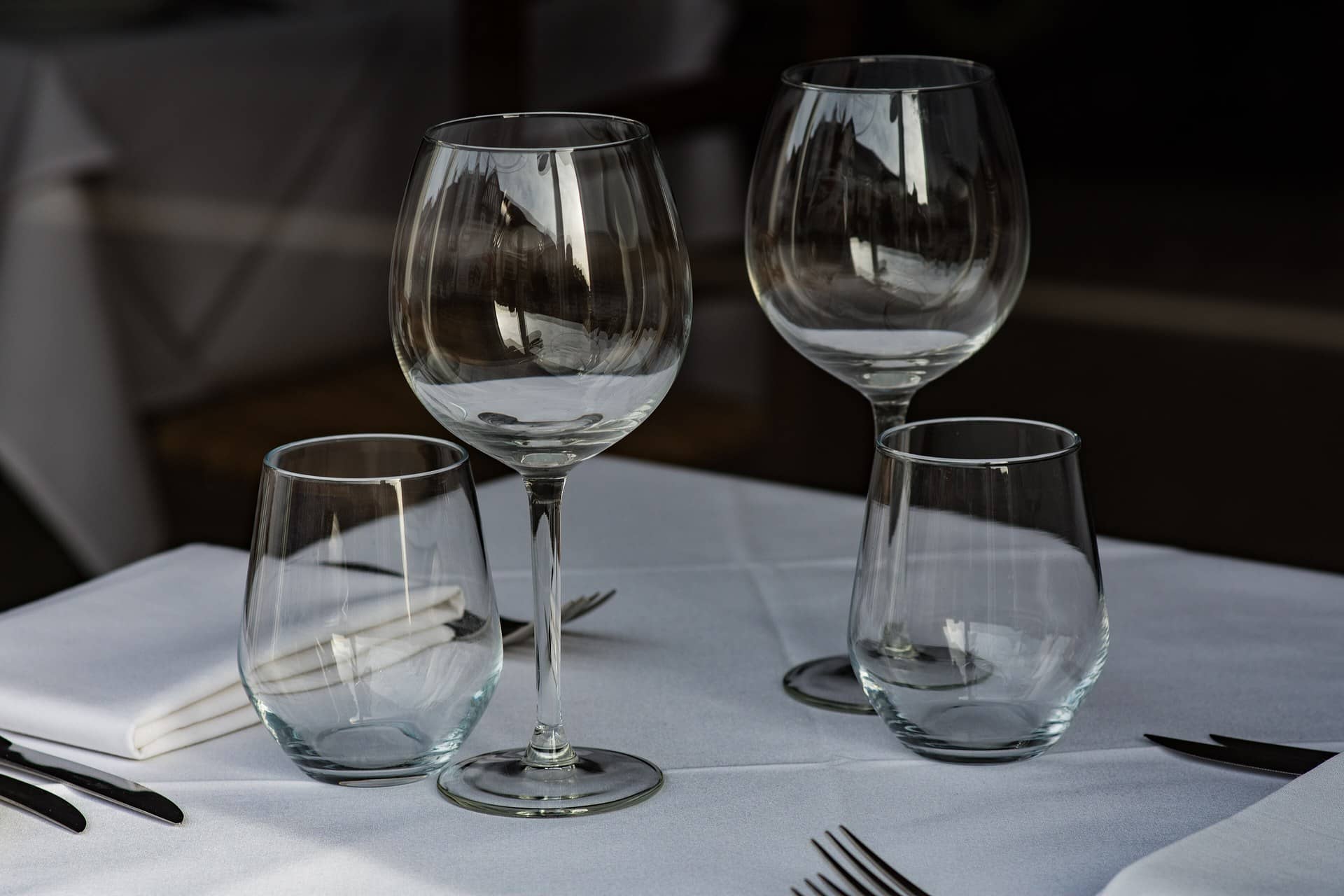 Empty wine glasses and tumblers on a white-clothed table with cutlery.