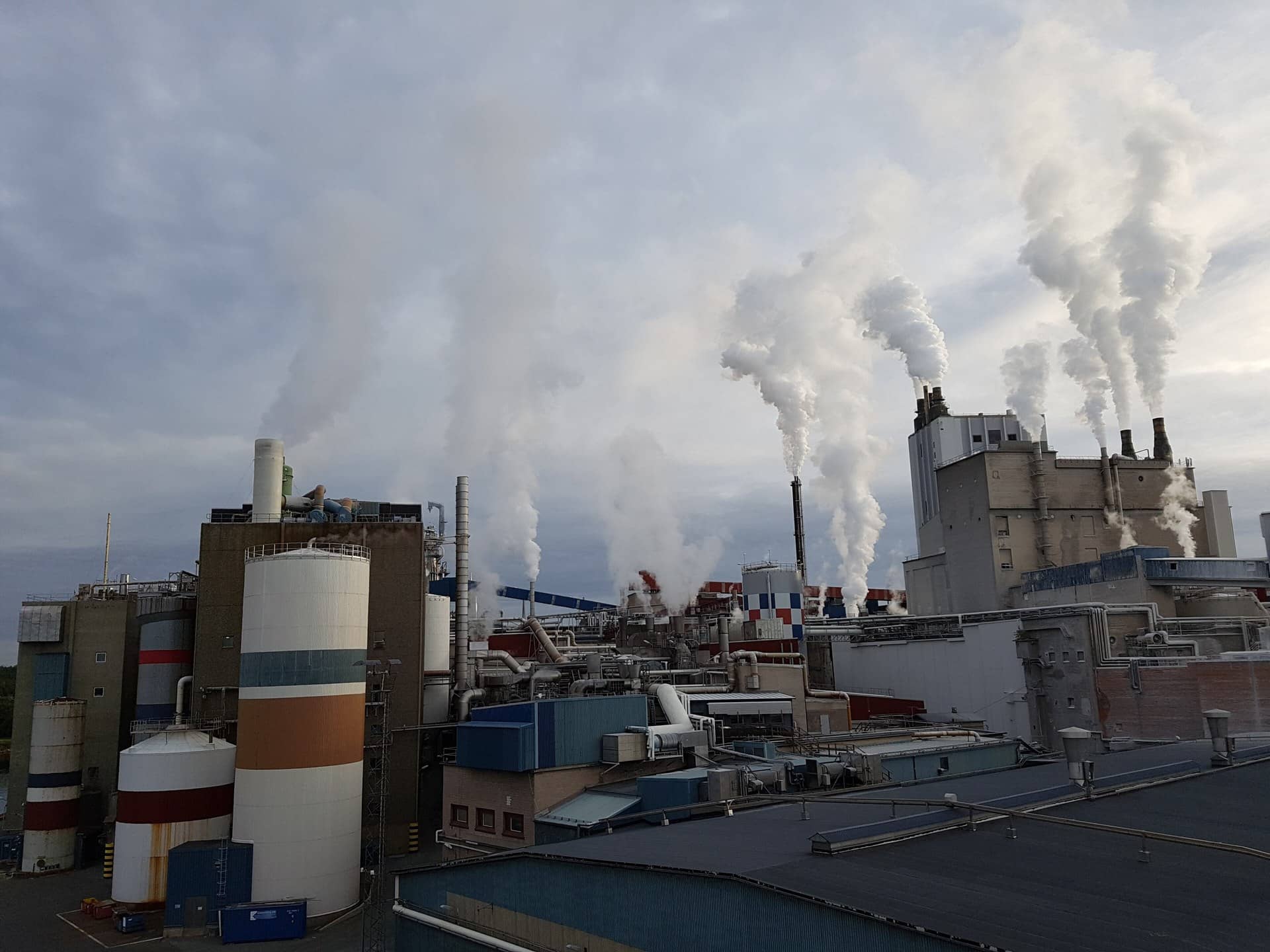 An industrial plant with storage silos and several white plumes rising into a cloudy sky.