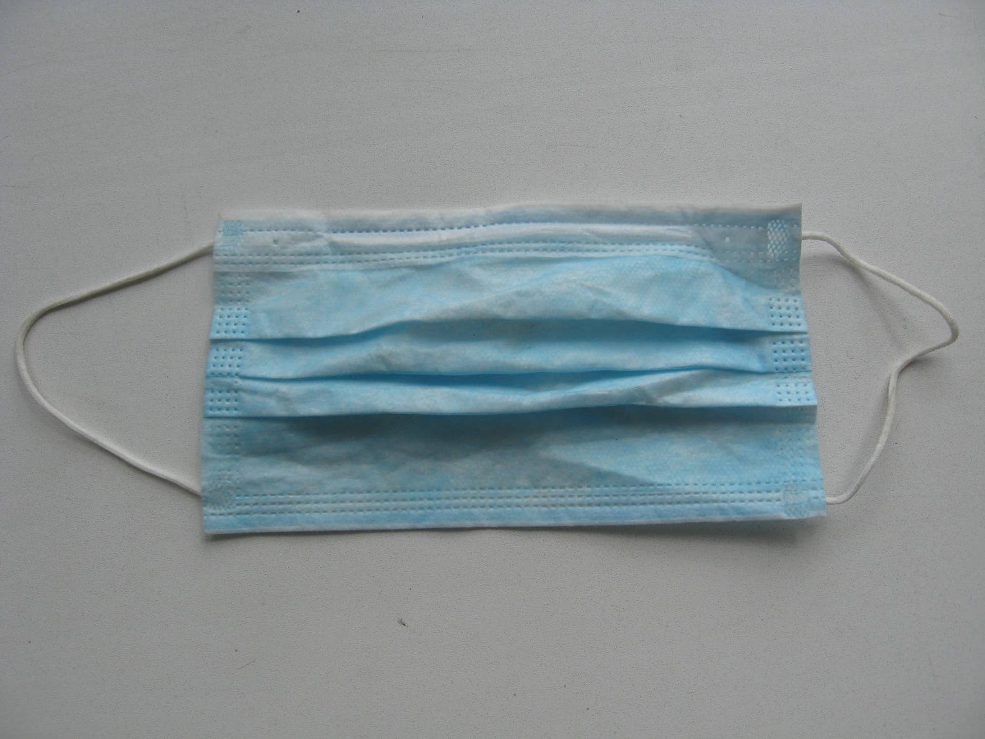 A pleated blue disposable surgical face mask with ear loops on a light grey surface.