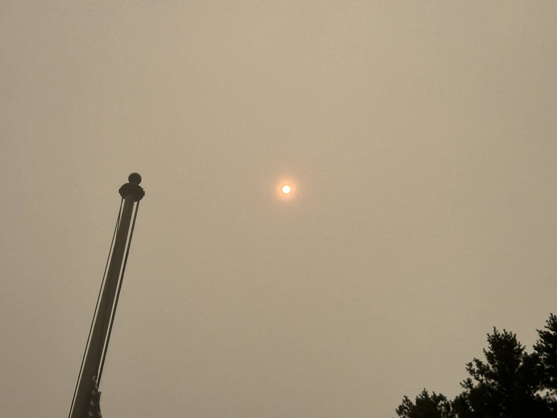 A small orange sun in a tan, smoke-hazed sky with a flagpole and tree tops at the edges.