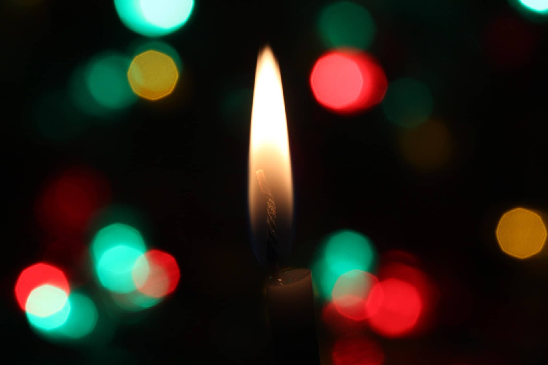 A single candle flame with soft coloured lights blurred behind it.