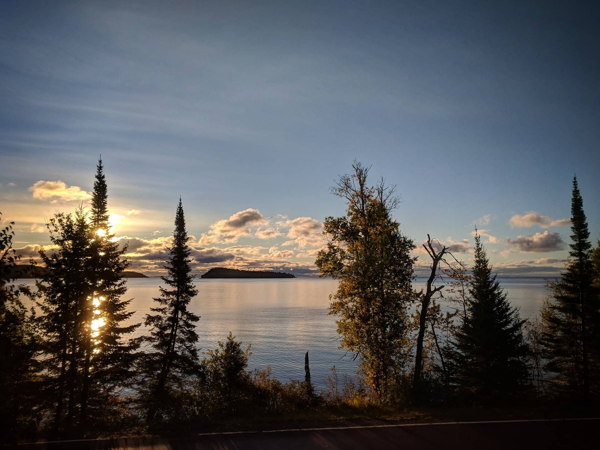 A calm lake with the sun low behind dark evergreen trees.