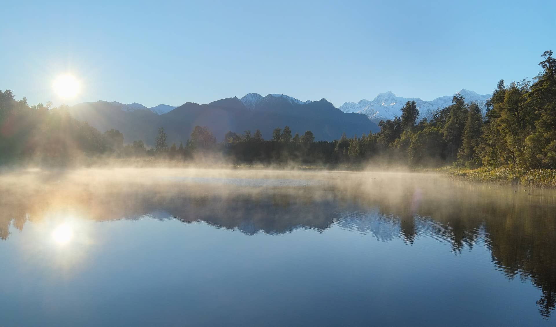 Mist over a still lake with snowy mountains and the low sun.