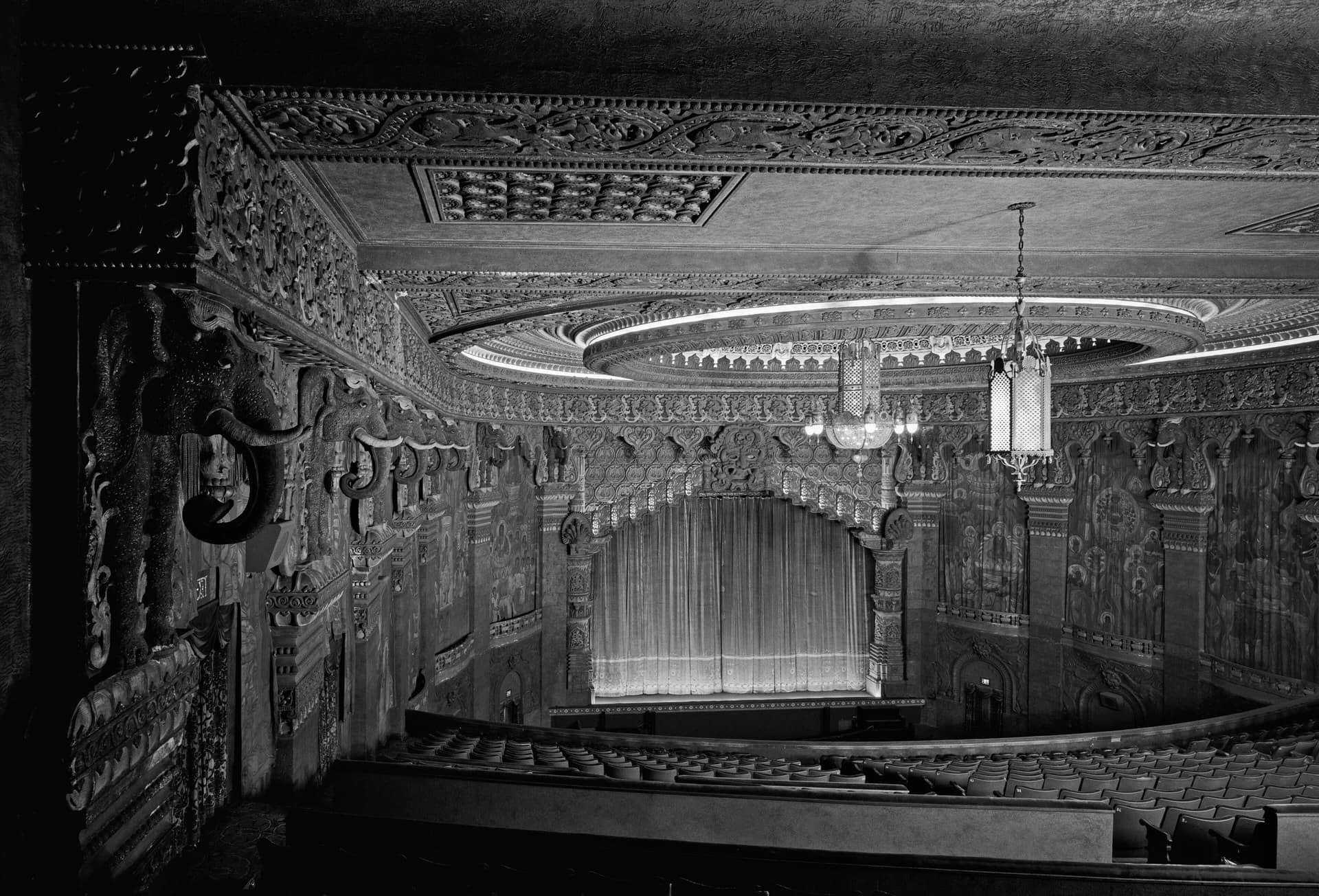 Black-and-white view of an ornate empty theatre auditorium from the balcony toward a curtained stage.