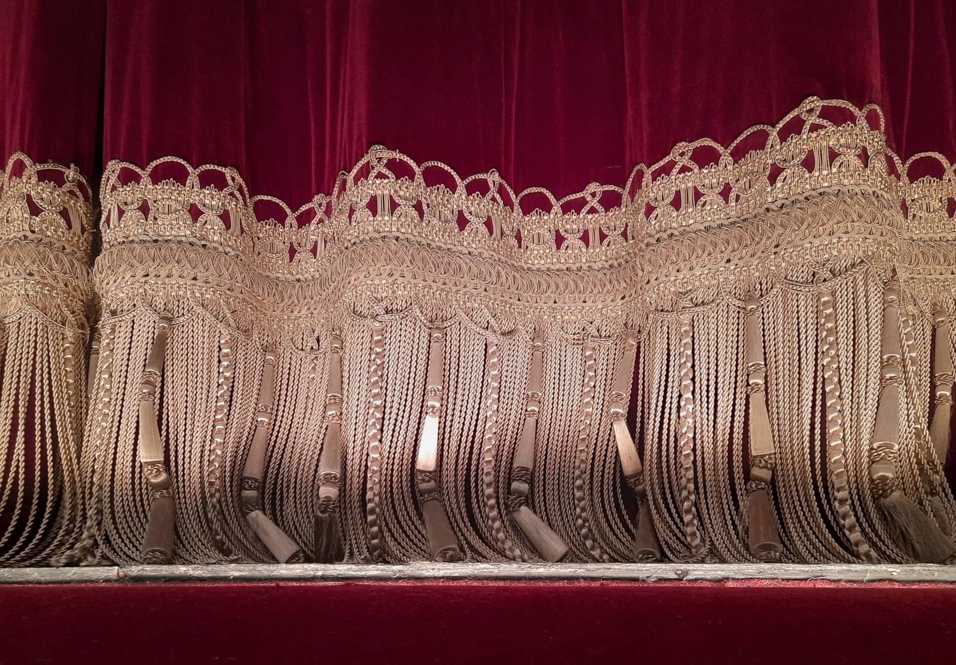 Close view of the tasselled gold fringe along the bottom of a red theatre curtain.