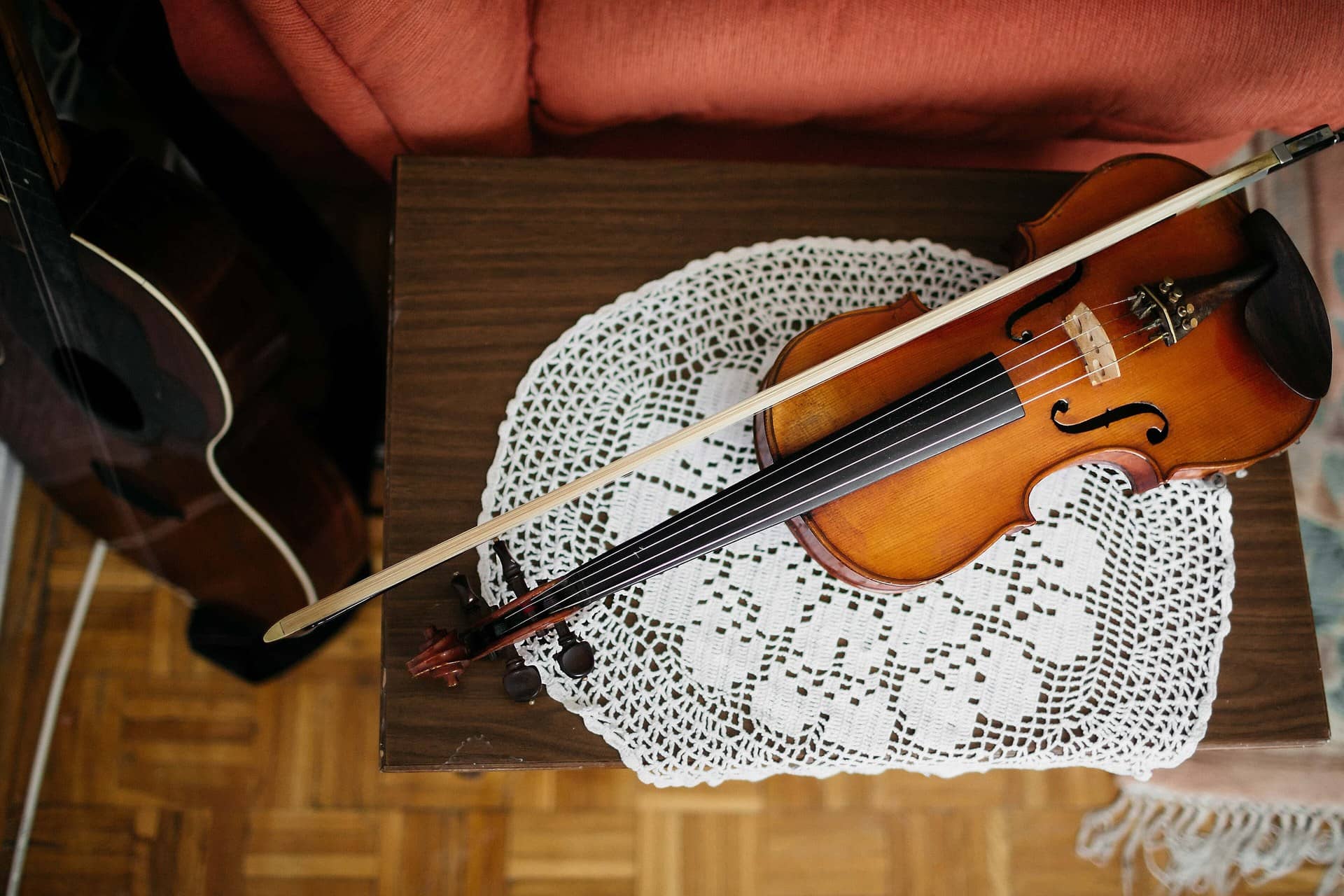 A violin and bow on a lace doily on a small wooden table, with part of a guitar at left.