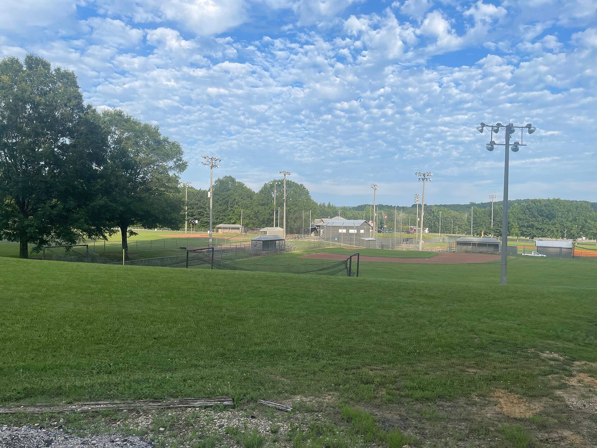 An empty baseball field with a dirt infield, floodlight poles and trees under a cloudy blue sky.
