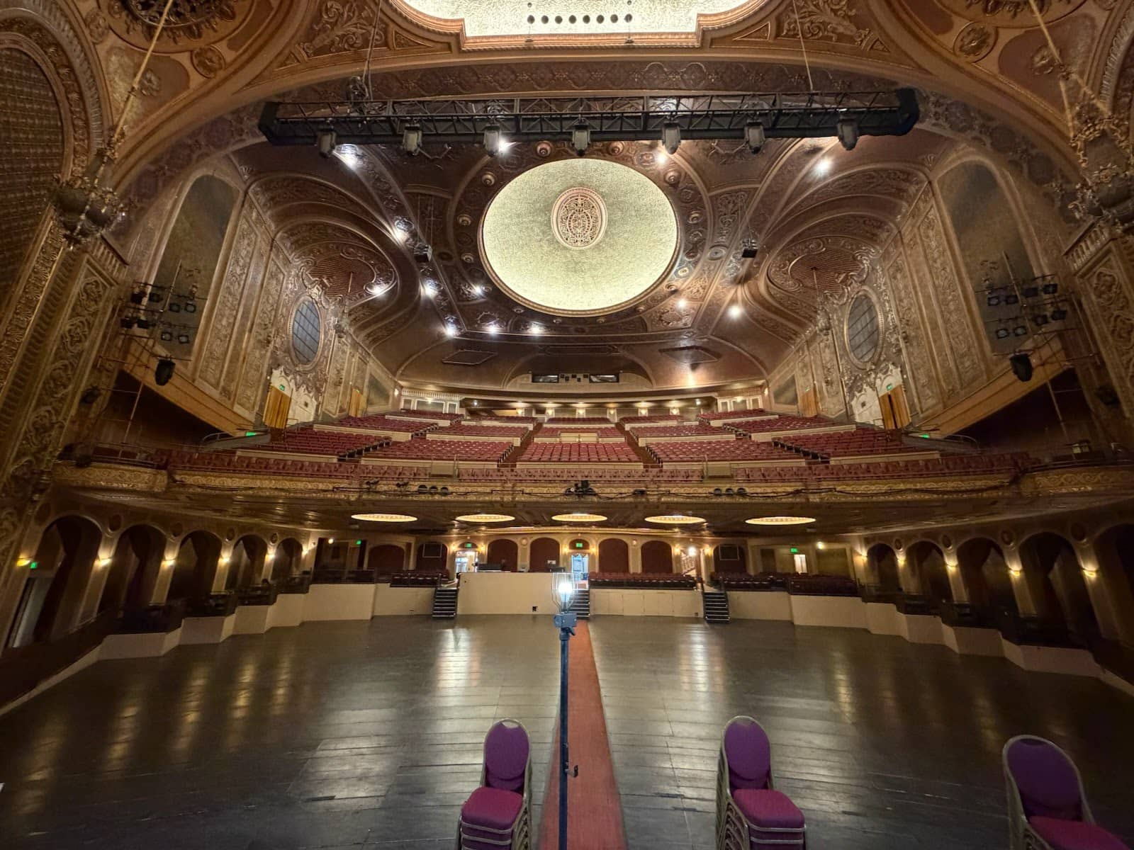 Wide view from an empty theatre stage toward ornate balconies and rows of red seats.