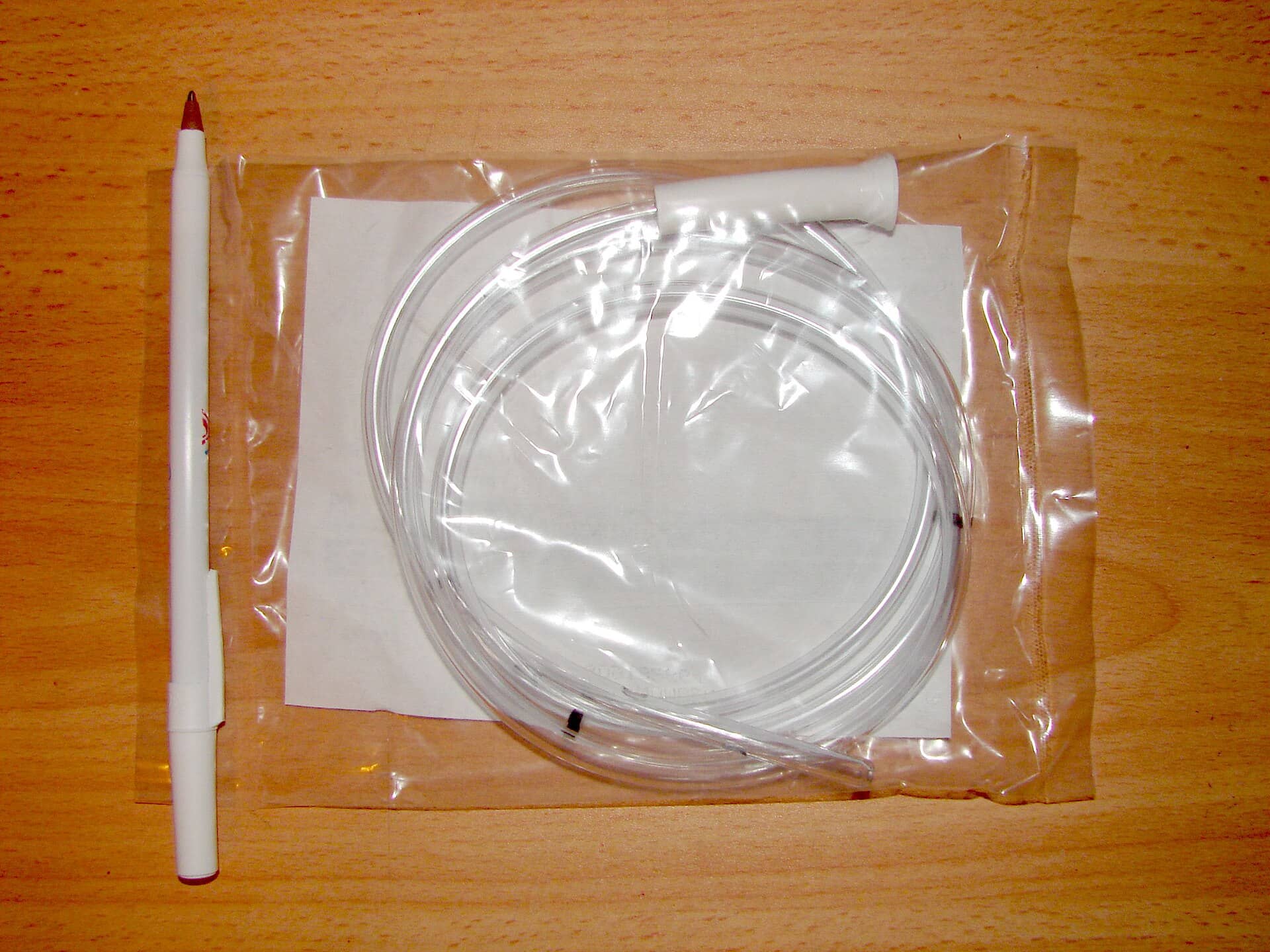 A coiled clear plastic tube in a sealed package on a wooden surface beside a pen.