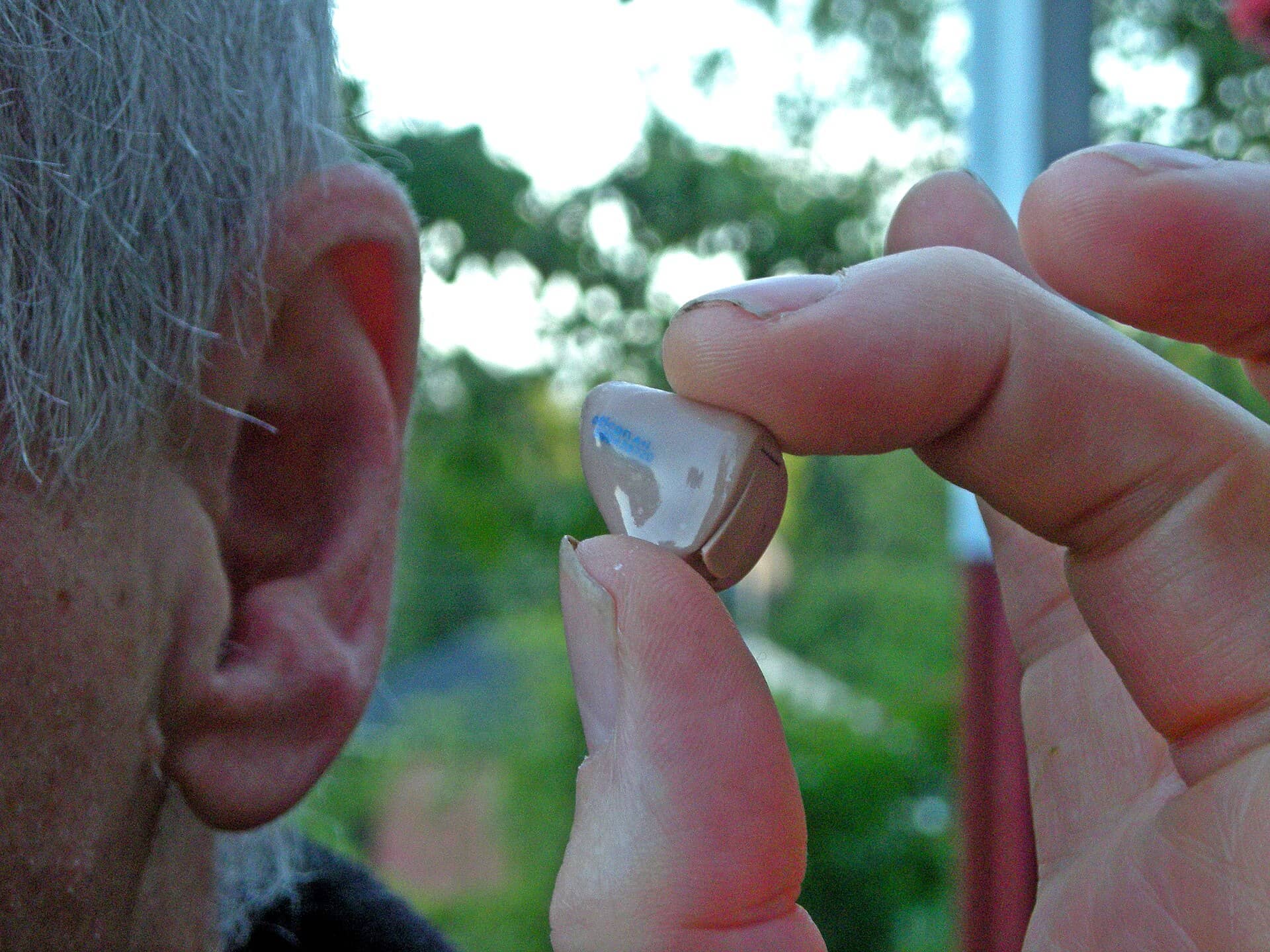 Fingers hold a small in-the-ear hearing aid beside a person's ear against a blurred green background.
