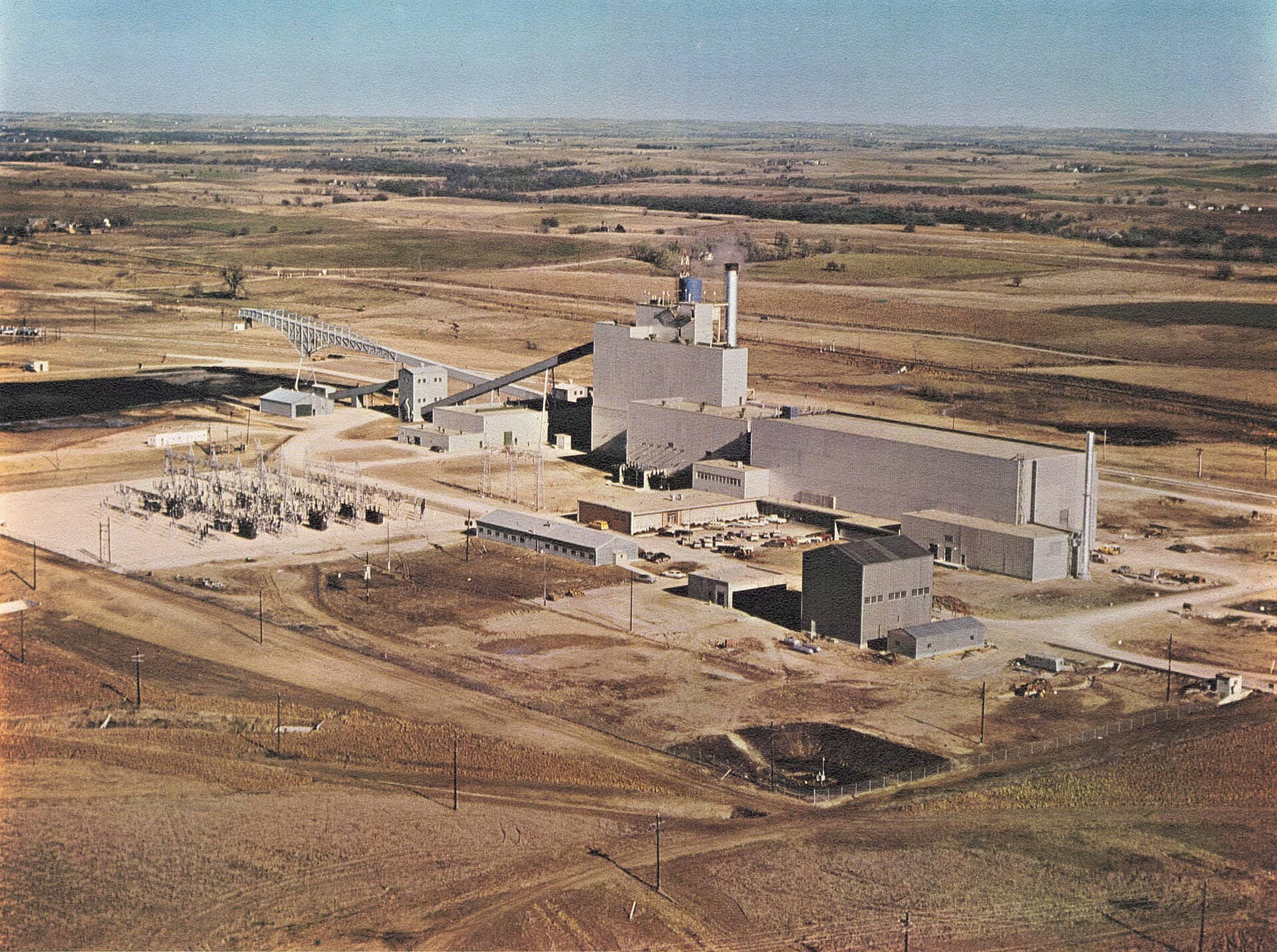An aerial view of an industrial power facility with large buildings, a smokestack and an electrical switchyard on open plains.
